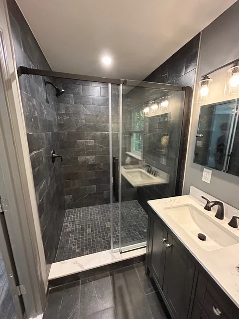 Dark slate tile shower with frameless glass door, dual white sinks, and vaulted ceiling