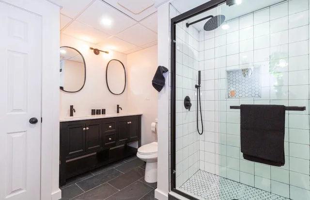 Bathroom with dark vanity, dual sinks, white subway tile shower, and dark tile flooring