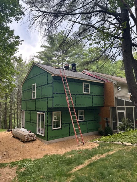 Two-story home with green siding, roofing work in progress, new windows, and staging ladder